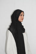 A woman wearing a black hijab with delicate lace trim along the edge, paired with a white abaya, shown in profile against a plain light background.